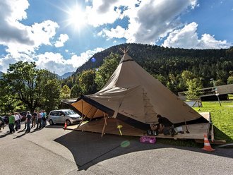 Ein Einzelhut-Tipi steht auf einem Parkplatz. Links neben dem Tipi steht eine Menschengruppe, im Hintergrund sind die Alpen zu erkennen.