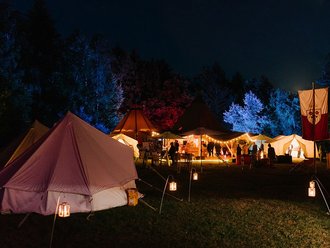Nachtszenario einer Tipi-Landschaft. Die Bäume im Hintergrund sind bunt beleuchtet, die Zelte strahlen, in warmen Farben.