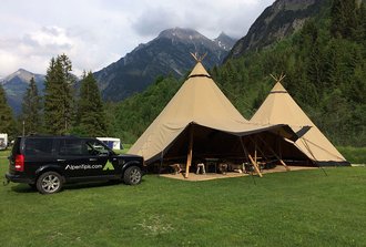 Auf einer Wiese in den Alpen steht ein 2er-Hut-Tipi. Neben dem Tipi steht ein schwarzes großes Auto von AlpenTipis.com.