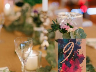 Auf dem Bild ist Deko zu erkennen, die auf einem Tisch steht. Es sind viele Blumen und Kerzen zu sehen. Im Vordergrund steht eine Karte mit bunten Farbklecksen, auf der die Zahl "9" steht.