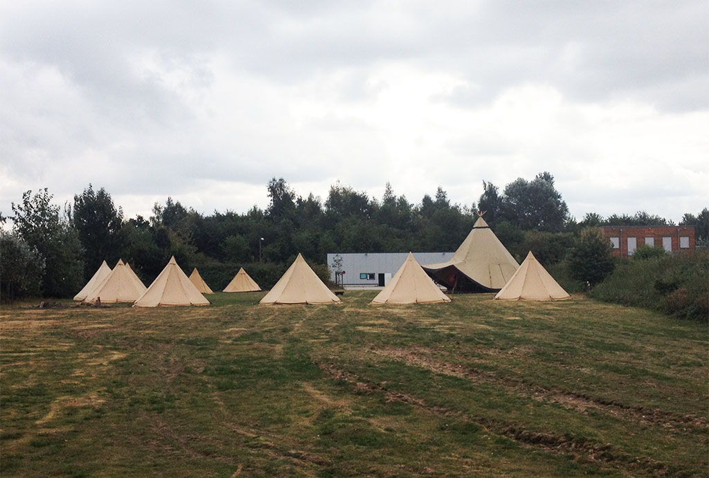 10 Einzeltipis stehen auf einer Wiesenfläche vor einem Industriegebiet.